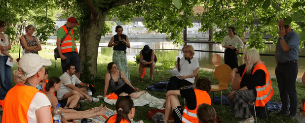 Ute Blohm-Hieber, Vorständin des Netzwerks Rechte der Natur, spricht beim Neckar Tribunal am Mikrofon. Teilnehmende in Warnwesten lauschen ihr unter einem Baum, dahinter ist der Neckar zu sehen.